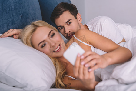 年轻人偷看微笑的女朋友使用智能手机在床上, 不信任的概念 