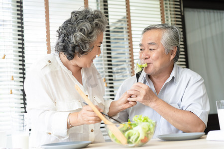 美丽的爱的亚洲老夫妇吃新鲜的蔬菜沙拉。妻子喂养丈夫, 并在家里的乐趣。温柔地看着对方.