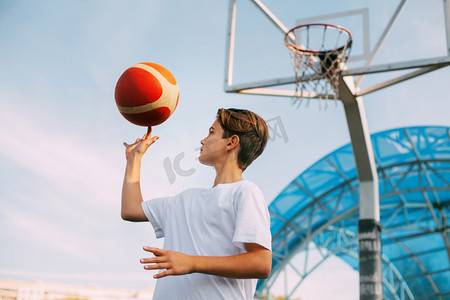 少年篮球摄影照片_一位身穿白色T恤的少年篮球运动员站在篮球场上，用手指捻着篮球。体育与健康生活方式的概念