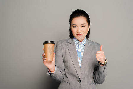 happy young businesswoman with paper cup showing thumb up on grey background