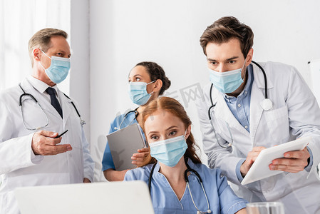 戴着医疗面罩的护士在医院多文化同事身边看着笔记本电脑，前景暗淡