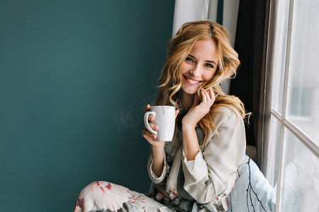 花冬摄影照片_欢快的金发女郎放松，坐在窗台上，拿着一杯咖啡，茶。她有长长的金发，美丽的微笑。穿漂亮的睡衣在花..