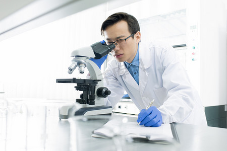 Asian medical scientist wearing white coat examining specimen using microscope, horizontal portrait