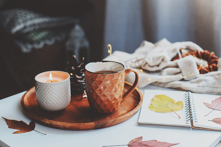 舒适的秋天或冬天的早晨在家。静止不动的生活细节与一杯茶，蜡烛，素描书与水族馆和温暖的毛衣。斯堪的纳维亚海格概念