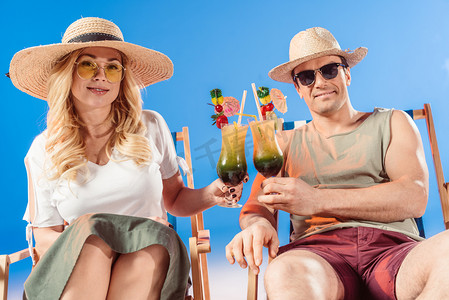 Young attractive couple toasting with cocktails resting in deck chairs on blue background