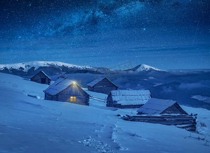 梦幻月光摄影照片_喀尔巴阡山脉的村庄由月光照亮, 山上的木制房屋覆盖着新鲜的积雪。梦幻般的银河在星空下。冬夜圣诞节.