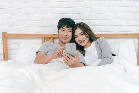 幸福的亚洲夫妇使用技术智能手机在床上的卧室在家里, 情侣和生活方式的概念,
