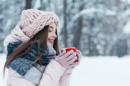 美丽的年轻女子在雪地公园手里拿着一杯热咖啡的侧面图