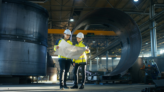 男性和女性工业工程师查看项目蓝图, 同时站在大型重工业制造工厂中间的管道部件周围
