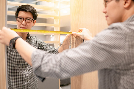 年轻的亚洲男子使用卷尺测量镜在现代木制橱柜的步行衣柜陈列室。家居装修家具。室内设计理念