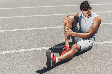 腿部受伤摄影照片_腿部受伤的年轻跑步者坐在跑道的地板上