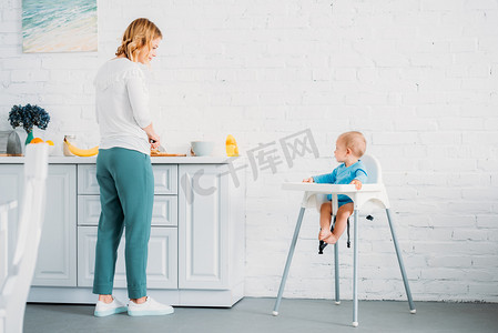 小孩子摄影照片_美丽的母亲为她的小孩子准备晚餐, 而他坐在厨房的喂养椅