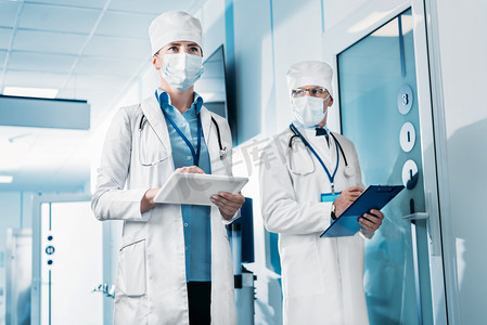 用数码片对医用口罩女医生的低角度观当她的男同事站在医院走廊的剪贴板后面 