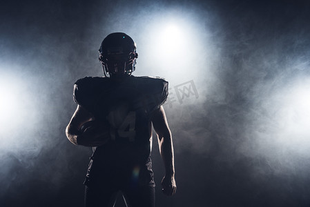 美国剪影摄影照片_装备美国橄榄球运动员的黑暗的剪影与球反对白色烟雾
