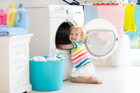 儿童在洗衣房内设有洗衣机或滚筒烘干机。帮助家庭琐事的孩子。现代家庭设备和洗涤洗涤剂在白色阳光的家。烘干机架清洗洗衣服. 