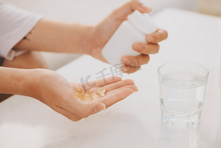 手与黄色胶囊欧米茄 3, 白色药丸的氨基葡萄糖和钙在塑料盒, 玻璃与水在木桌上, 顶部视图.