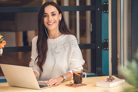 有吸引力的年轻女性自由职业者在耳机工作在膝上型电脑和看照相机在桌与酒在咖啡馆里 