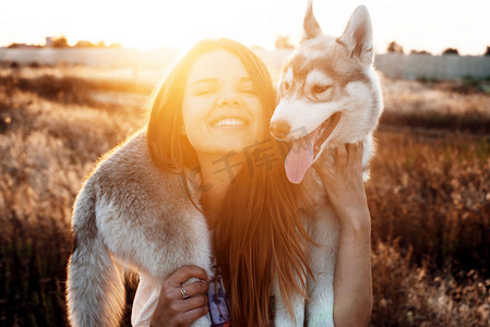 年轻的白种的女性，在夕阳下玩弄她在外地的西伯利亚哈士奇犬小狗。快乐的微笑女孩玩的小狗在户外在美丽的光