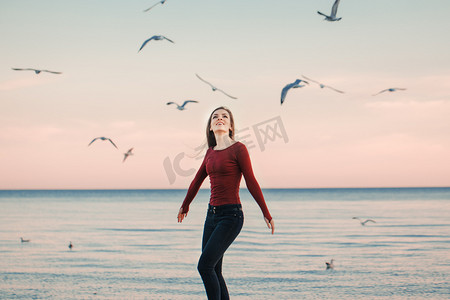 电影龙摄影照片_肖像的幸福的微笑笑兴奋高加索运行跳海鸥鸟群当中日秋高气爽的秋季户外在岸滩上在日落，生活方式自然情感的牛仔裤的年轻女子