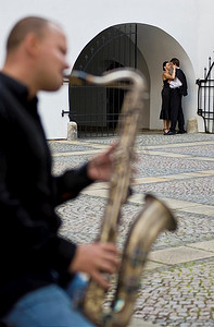 情侣浪漫背景摄影照片_一位街头音乐家吹着萨克斯管，一对浪漫情侣在背景中接吻的选择性对焦镜头