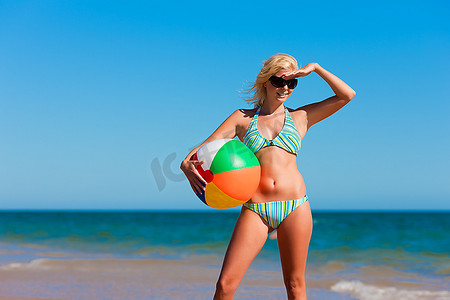 一位穿着比基尼的迷人女子站在蓝天下的海滩上，用手捂住眼睛