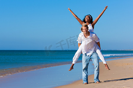 恋爱中的情侣--一名白人男子在海滩上的蓝天下背着他的非裔美国女人