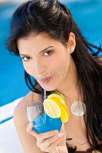 美丽的拉美裔年轻女子在蓝色游泳池边喝着鸡尾酒