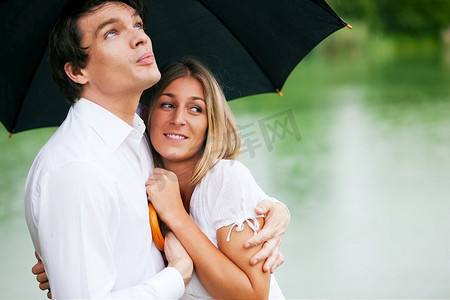 一对情侣(一男一女)在夏天的湖边撑着雨伞，他抱着他的女孩，为她遮挡雨滴