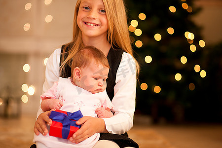 子代摄影照片_两个姐妹--一个还是个婴儿--拿着礼物站在圣诞树前