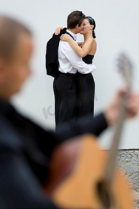 一对相爱的年轻夫妇拥抱在一起，一位街头音乐家在前景中弹着吉他(模糊)。