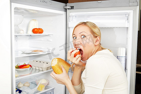 贪婪的女人靠着开着的冰箱吃饭