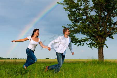 彩虹朋友摄影照片_夏天，幸福的情侣在草地上奔跑；背景是一道彩虹