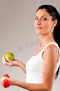一位运动女性拿着灰色背景上的红色杠铃和绿色苹果