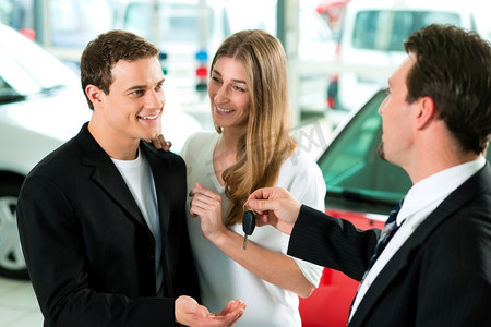 汽车站摄影照片_在一家汽车经销商的销售情况下，经销商正在把汽车钥匙递给一对年轻夫妇，他们很兴奋，汽车站在背景中