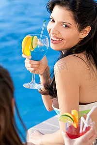 一位年轻漂亮的拉美裔女子在蓝色游泳池边喝着鸡尾酒，她的朋友在前景中大笑着。