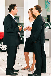 在一家汽车经销商的销售情况下，经销商正在与一对年轻夫妇交谈，背景中站着几辆车