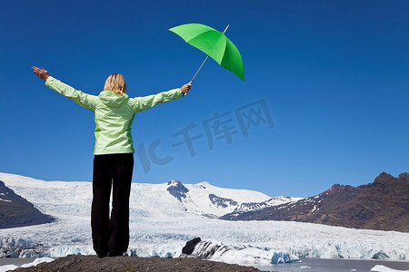 环境概念照片，一名金发女子穿着绿色衣服，手持绿色雨伞，站在融化的冰川前张开双臂