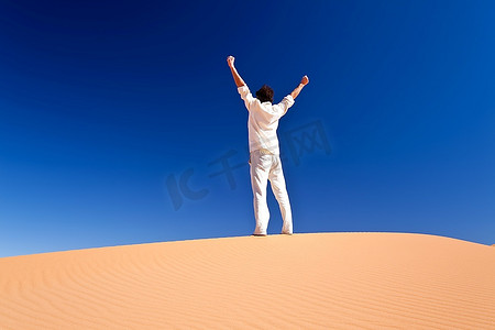 背影白人摄影照片_一名成年白人男子站在沙丘上举起双臂的背影。Erg Chebbi，Maroc