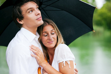 一对情侣(一男一女)在夏天的湖边撑着雨伞，他抱着他的女孩，为她遮挡雨滴