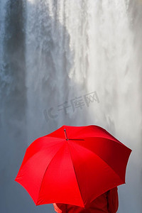 一名身穿红色夹克、手持红色雨伞的女子站在瀑布前。在冰岛斯科加福斯外景拍摄。