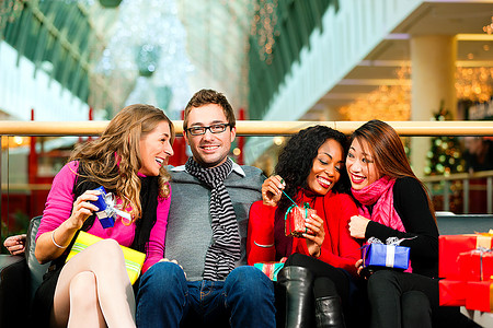 由四个人组成的多元化团体--白人、黑人和亚洲人--坐在一家购物中心里，拿着圣诞礼物和袋子