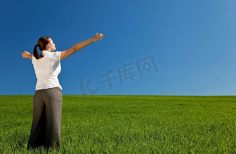 一位美丽的年轻女子，张开双臂赞美或庆祝，站在绿色的田野上，面朝蓝天中的光