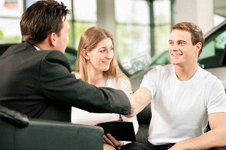在一家汽车经销商的销售情况下，经销商正在与一对年轻夫妇握手，他们想把车放在背景中