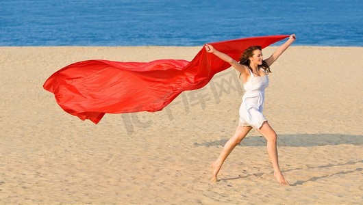 一个性感和美丽的年轻黑发女子沿着金色的海滩与红色材料的长度和深蓝色的大海在她身后运行。