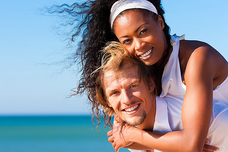 恋爱中的情侣--一名白人男子在海滩上的蓝天下背着他的非裔美国女人