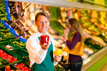 超市里的售货员在菜架上；在背景中，一名妇女正在挑选蔬菜