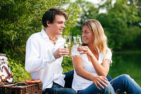 一对坐在湖边用白葡萄酒碰杯野餐的情侣