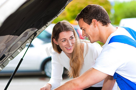 一名妇女在停车场与一名汽车修理工交谈，两人都站在汽车旁边