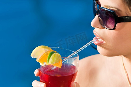 令人惊叹的美丽拉美裔年轻女子在蓝色游泳池边喝着红色鸡尾酒