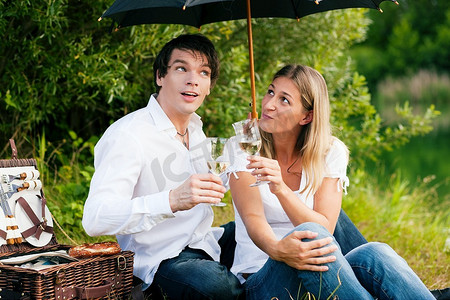 一对相爱的情侣坐在湖边喝着酒野餐--天开始下雨了，但他们不在乎